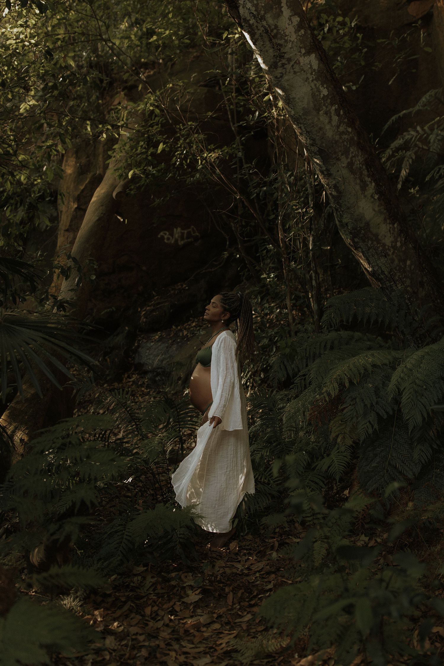 Pregnant woman in white dress standing among lush ferns and trees in a dark forest.