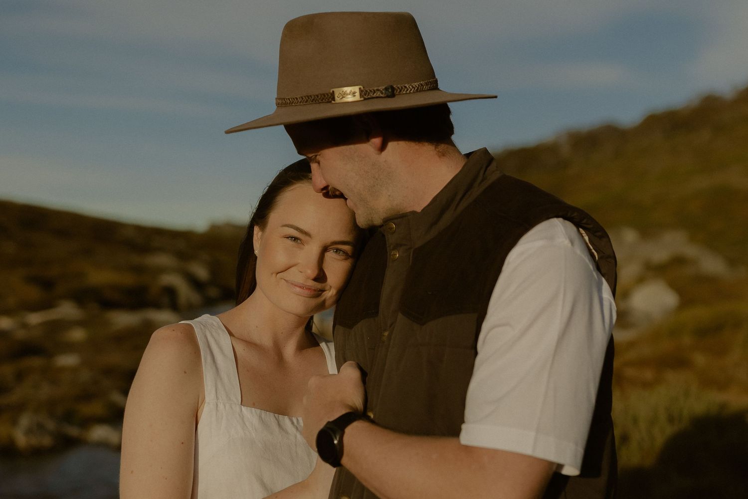 A couple in western attire embraces on a scenic hillside during golden hour.