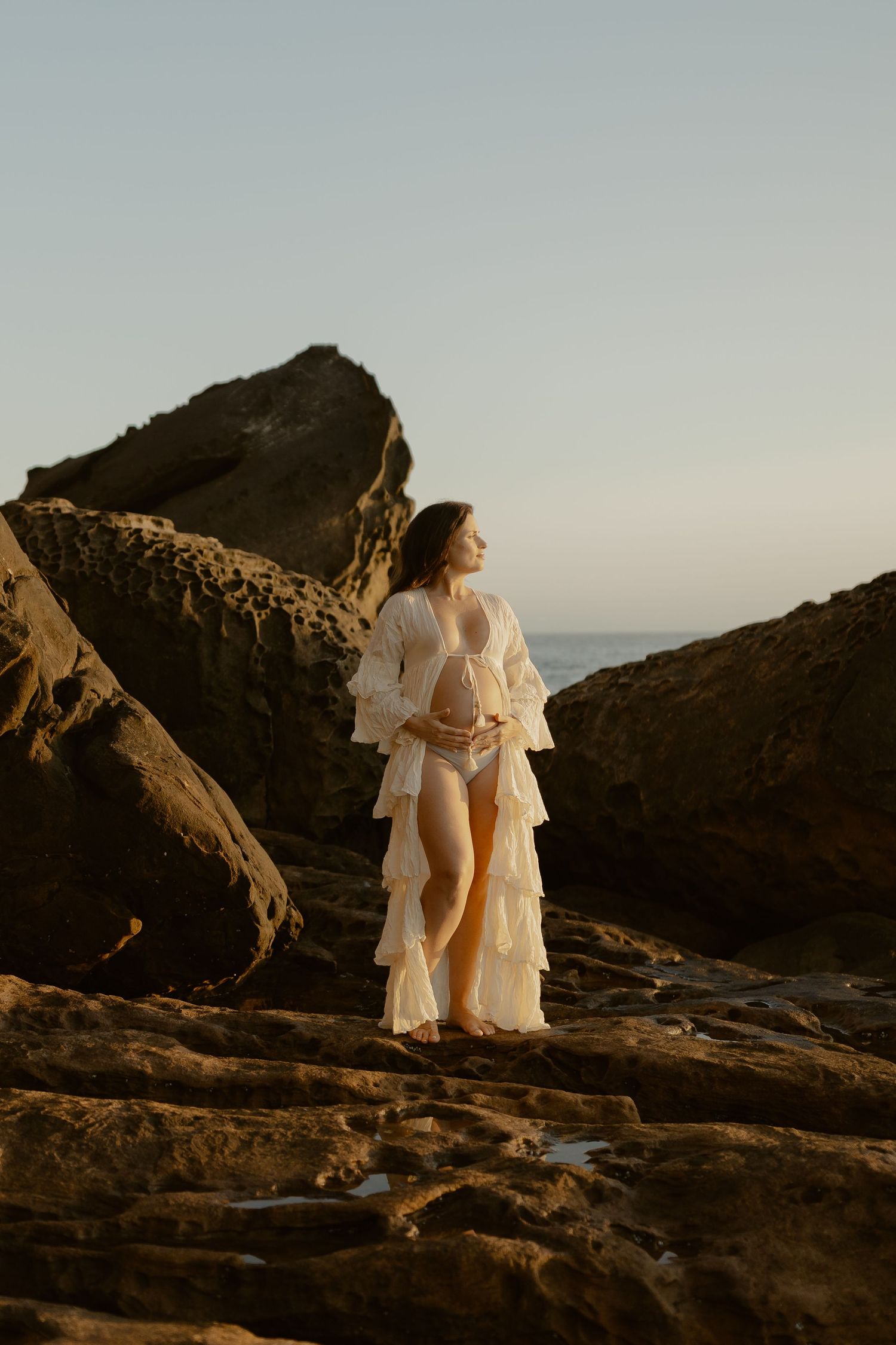 Two figures in flowing white dresses stand together on rocky coastal cliffs at sunset.