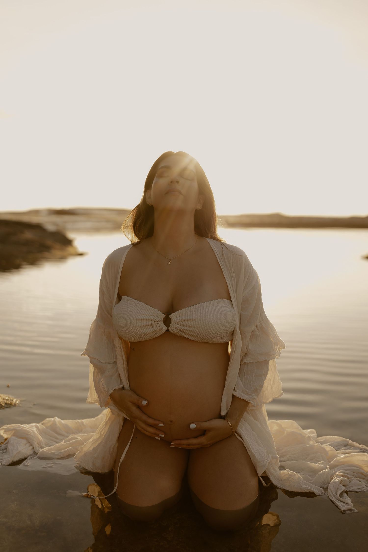 A silhouetted figure in white swimwear sits by a serene lake during golden hour, creating a peaceful maternity moment.