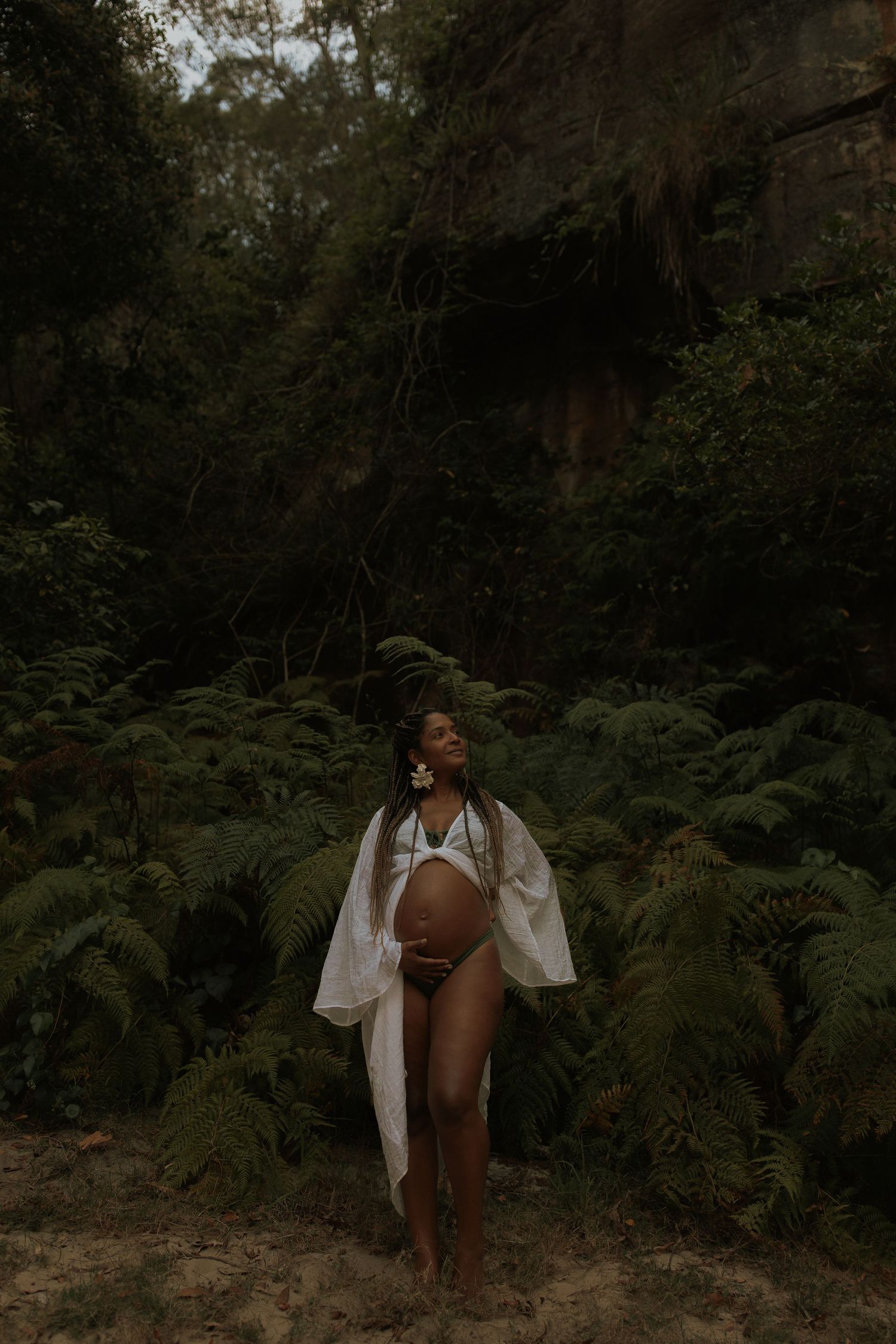 A silhouette poses gracefully among lush ferns in a moody, forest setting wearing white flowing fabric.