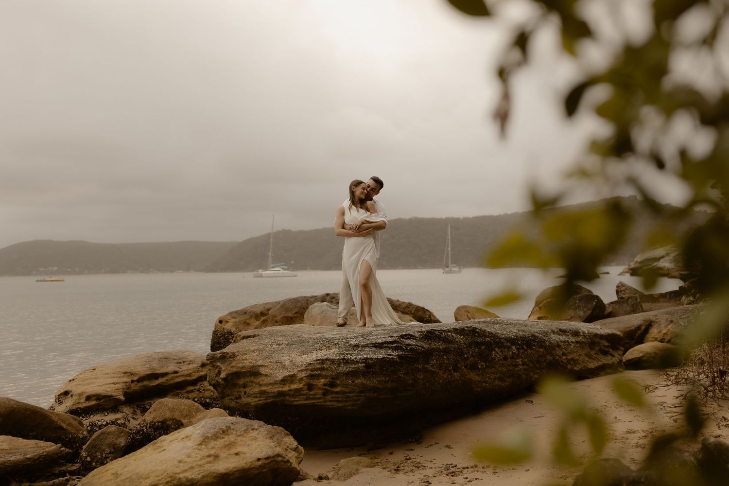 A couple embraces on a rocky cliff overlooking a misty coastal landscape at sunset.