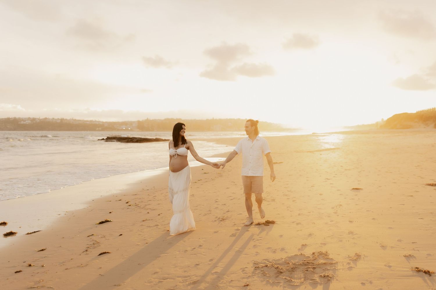Two people walk along a sandy beach at sunset while holding hands and leaving footprints in their wake.