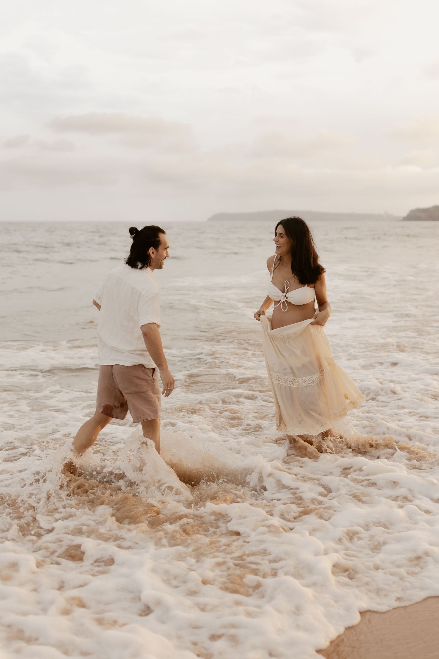 Romantic couple playing in ocean waves during sunset beach photoshoot.