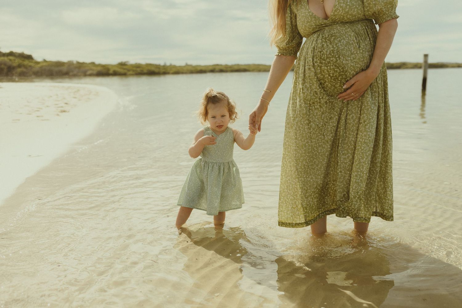 A mother and child walk together along a sandy beach shoreline on a sunny day.