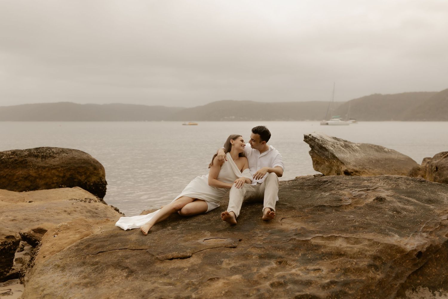 A couple in wedding attire shares romantic moments on a rocky coastal cliff overlooking the ocean at sunset.