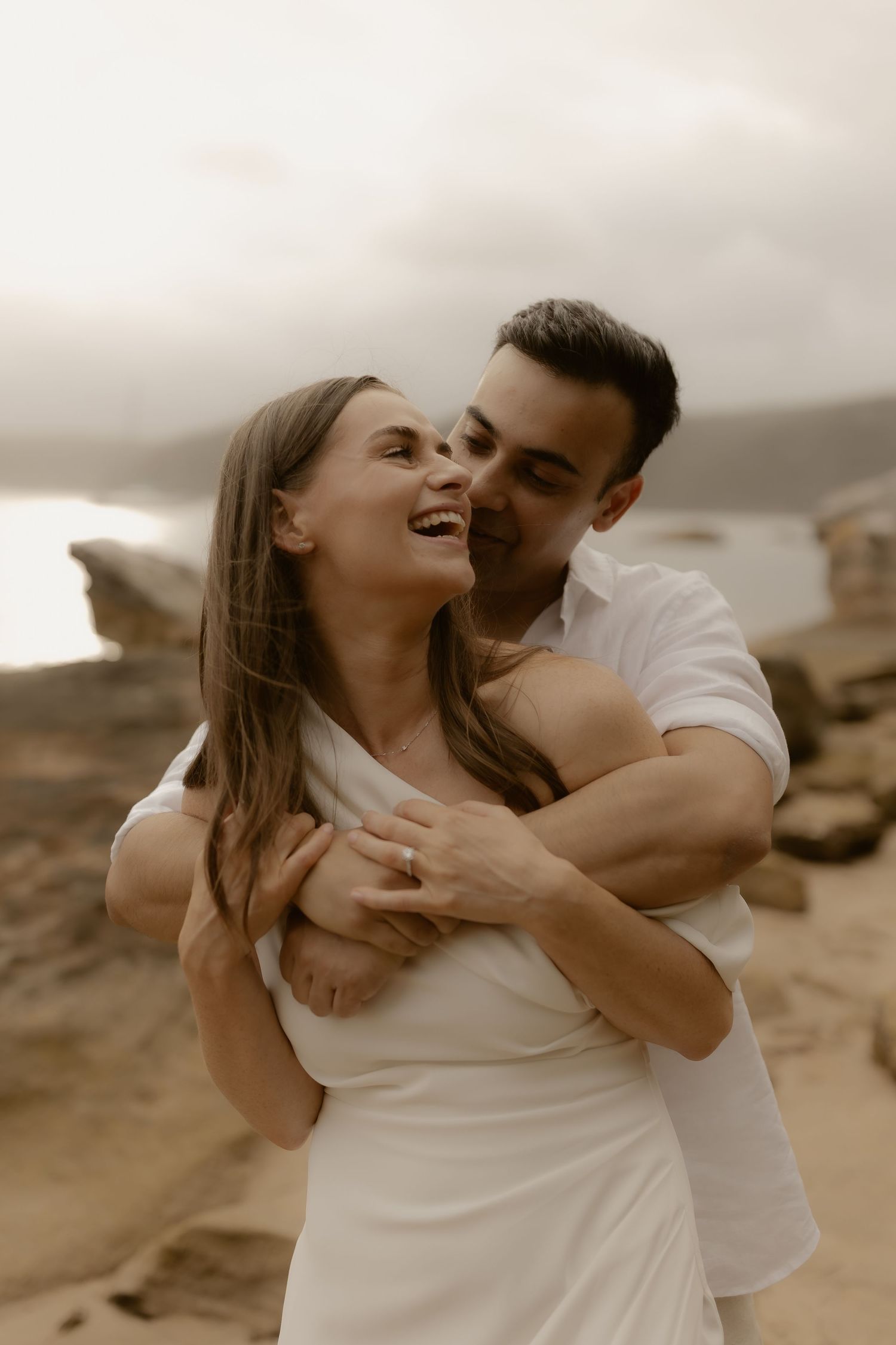 A romantic couple in white embraces and laughs together on a cloudy beach.