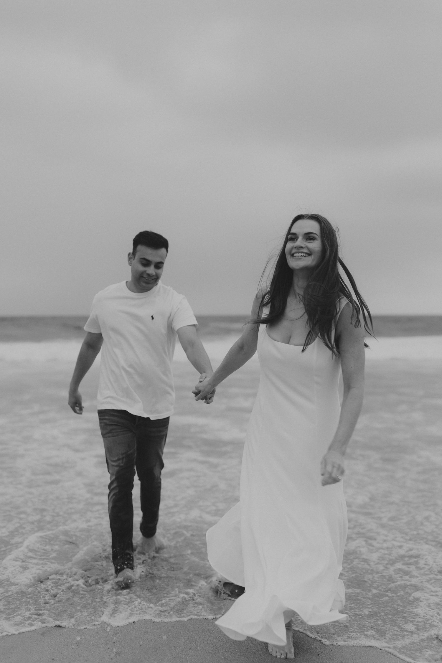 Couple running and playing in ocean waves wearing white and casual attire in black and white photo.