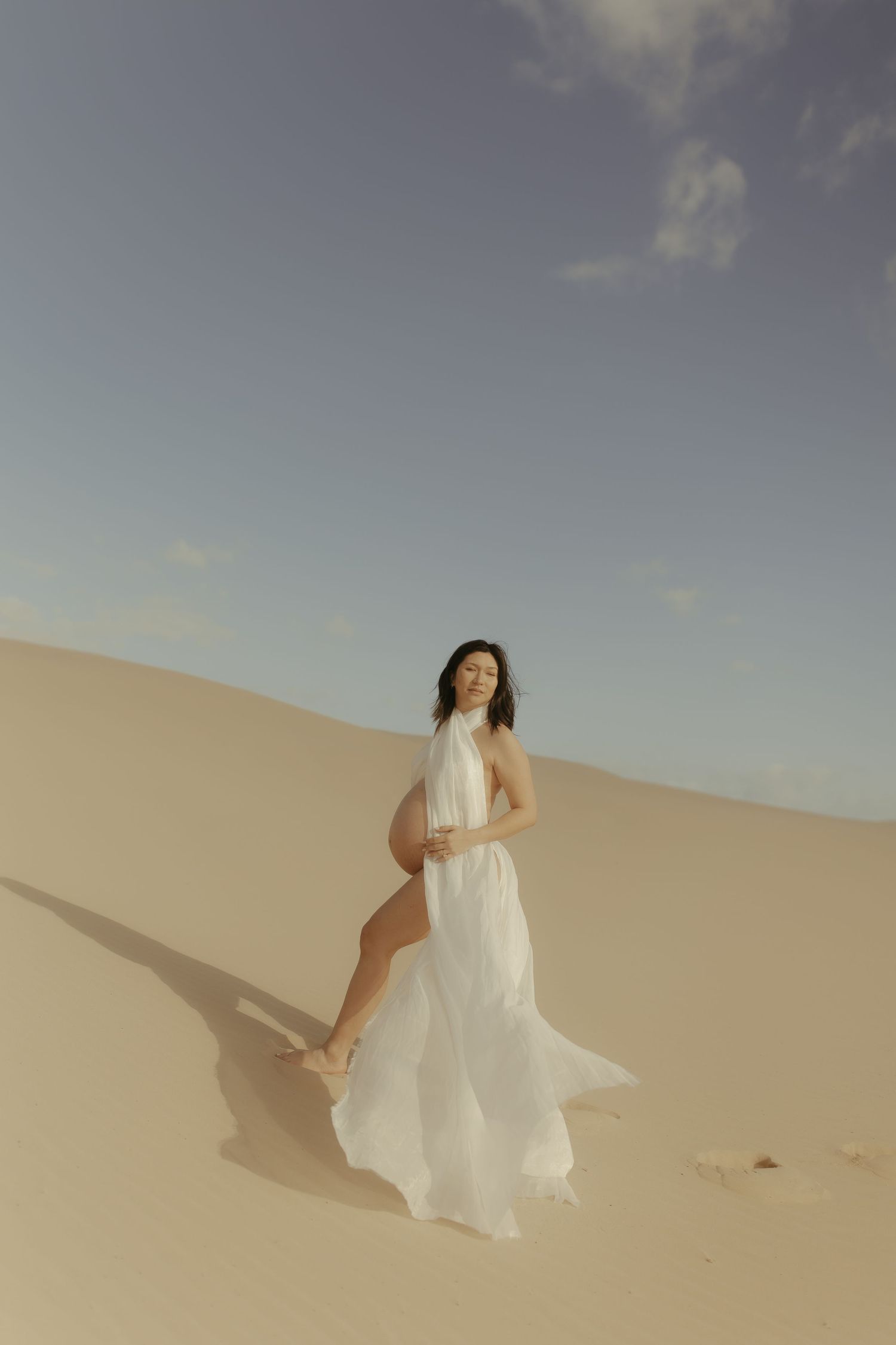 A person in a flowing white dress dances gracefully across golden sand dunes under a soft cloudy sky.
