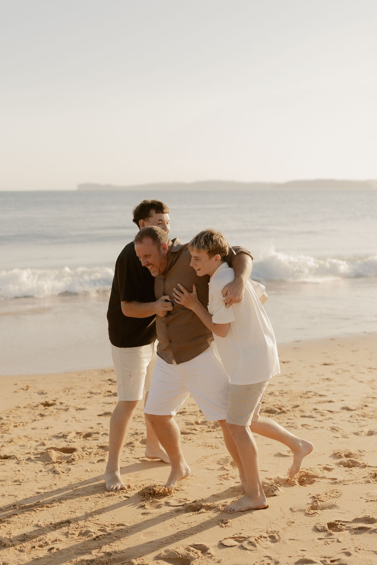 Three people playfully running and laughing together on a sandy beach at sunset.