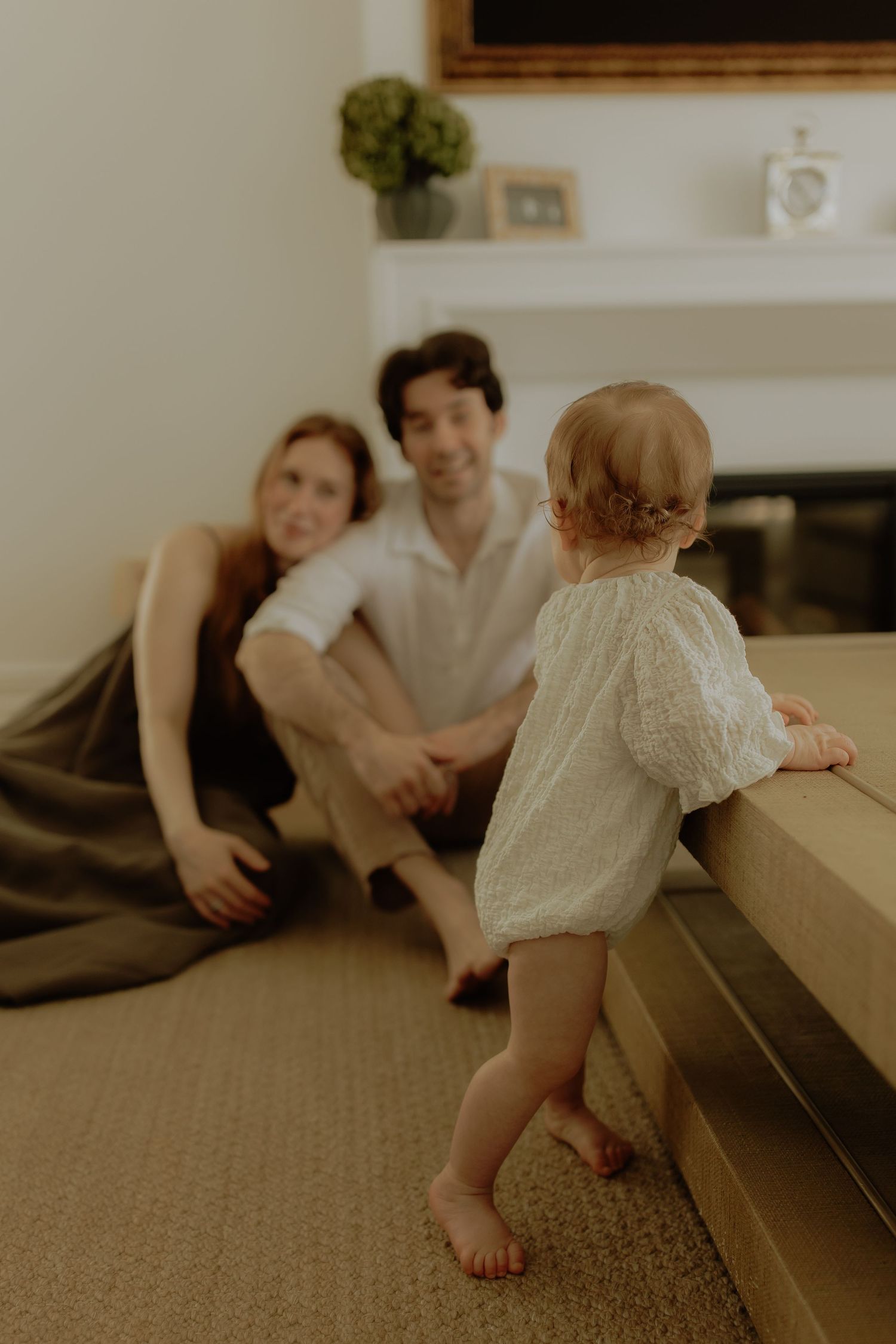 A baby in a white dress stands at a couch while parents sit on the floor smiling in a cozy living room.