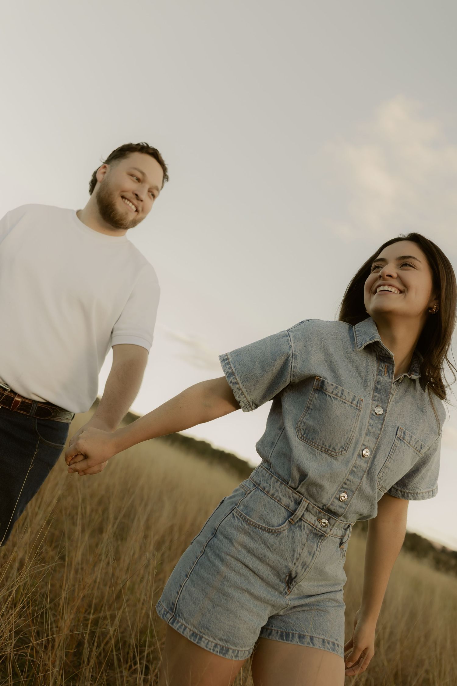 Happy couple holding hands and laughing while walking through a golden grass field at dusk.