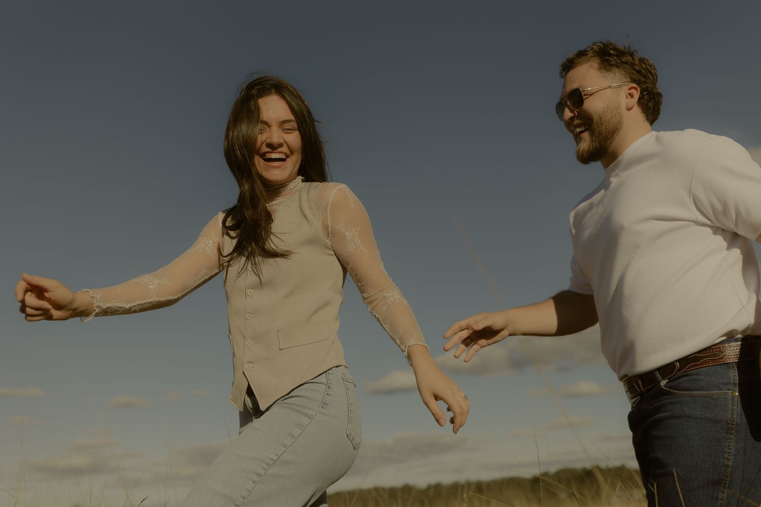 A joyful couple laughs and dances outdoors under a blue sky, enjoying a carefree moment together.