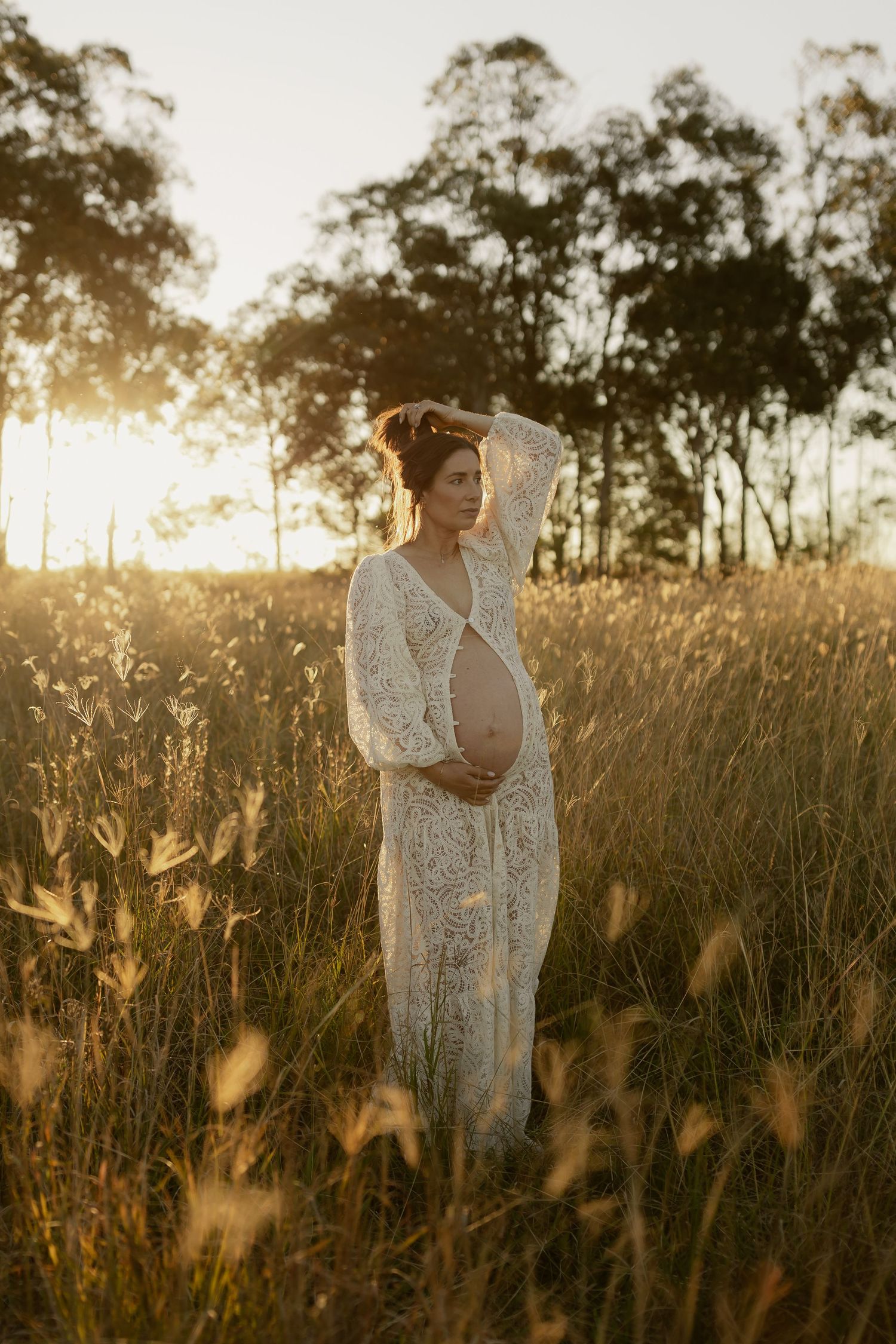 Pregnant woman in white lace dress cradling belly in sunlit golden grass field at sunset.