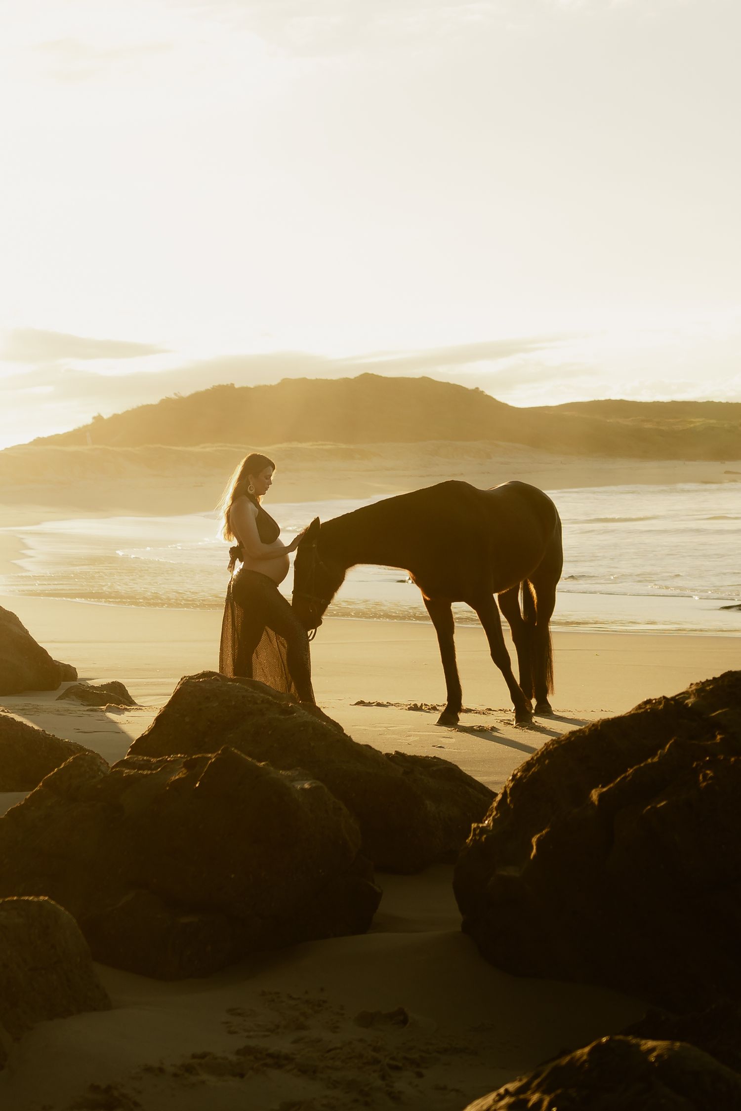 Woman standing with a dark horse on a golden sunset beach, with rocky shores and ocean hills in the background.