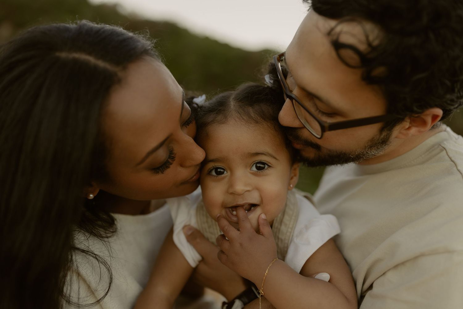 Two parents lovingly kissing their smiling baby on the cheeks outdoors in a warm, candid family portrait.