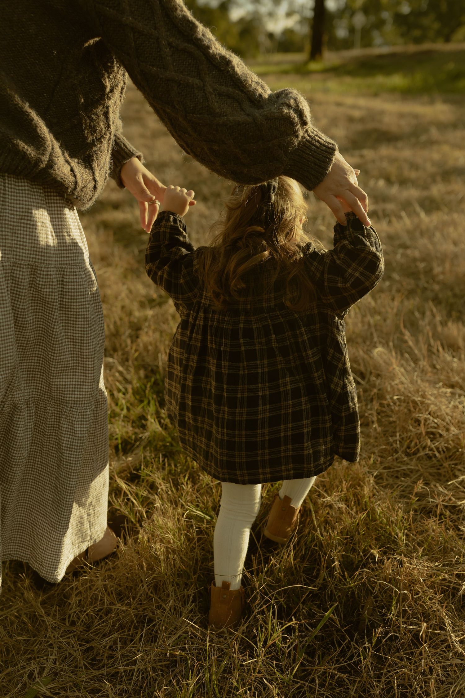 A young girl in a plaid dress holds her mother's hands while walking through a golden sunlit field in autumn.