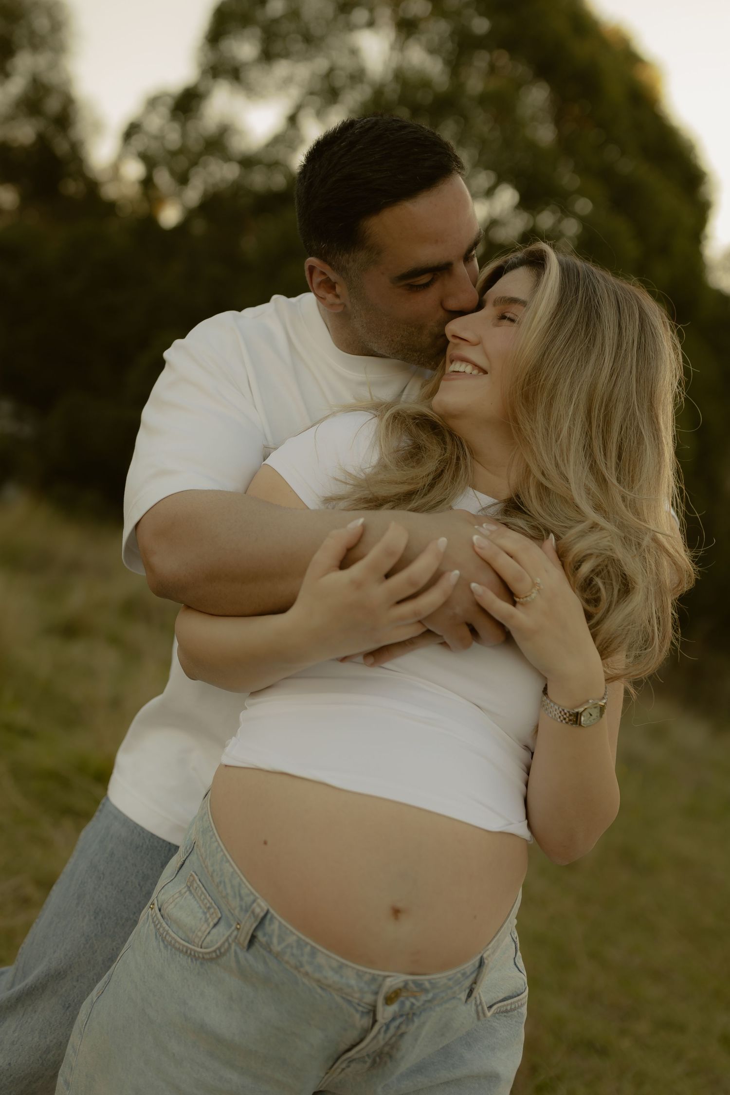 Man embracing pregnant woman from behind, kissing her cheek in a sunlit park during maternity photoshoot.