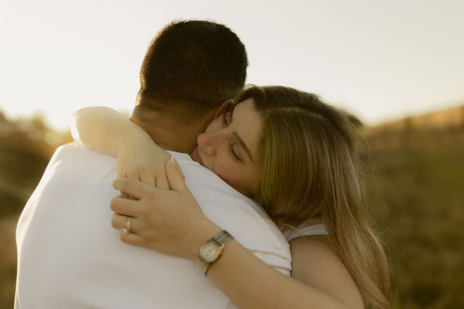 A couple shares a tender embrace at golden hour outdoors, with the woman wearing a watch and white outfit.