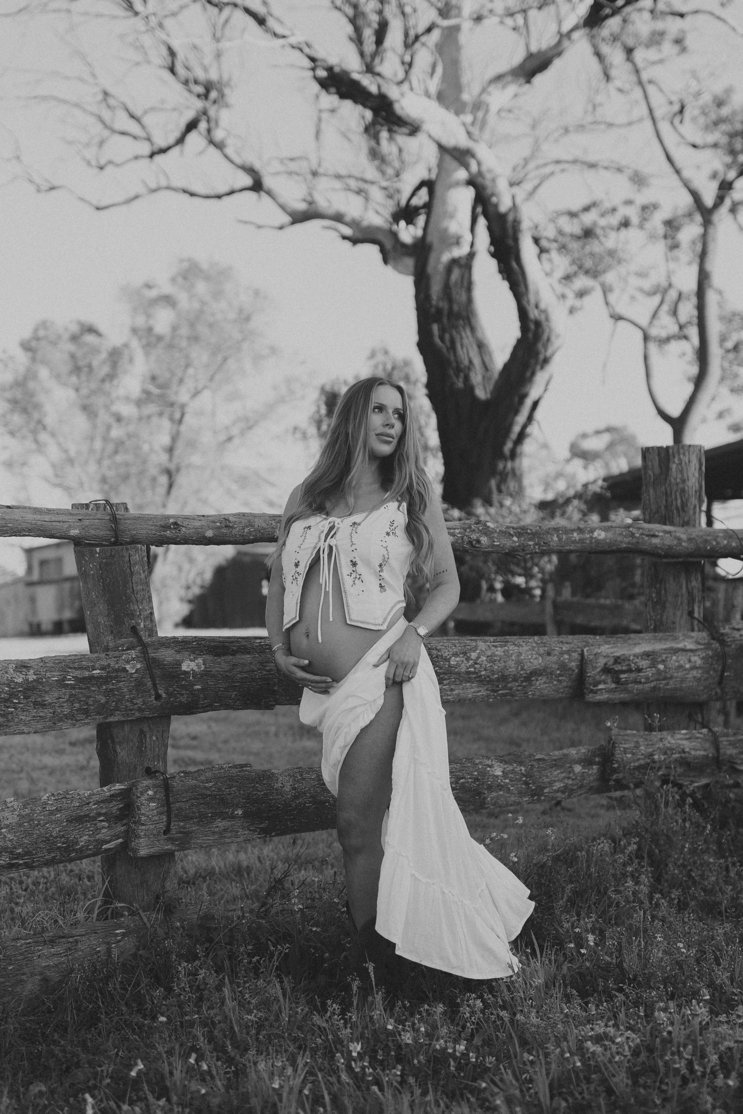Woman in white maxi dress leans against rustic wooden fence with large tree backdrop in black and white photo.