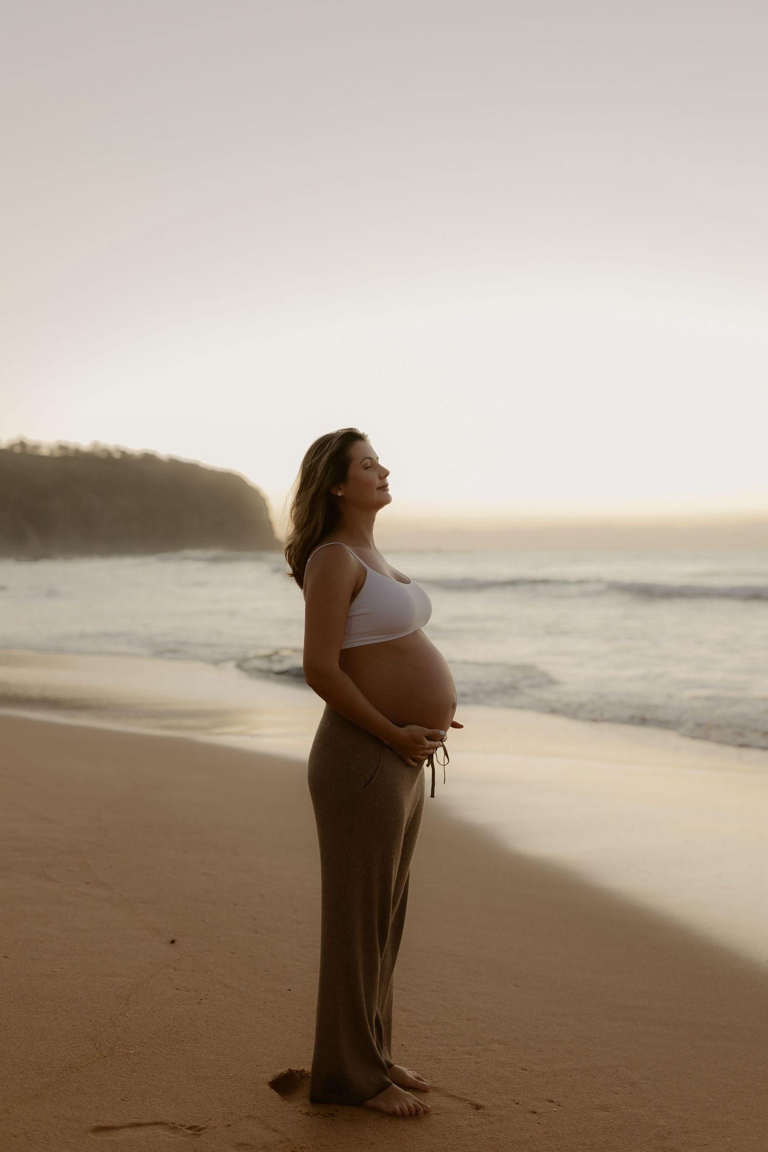 Pregnant woman in white crop top and olive pants cradling her belly on a sandy beach at sunset.