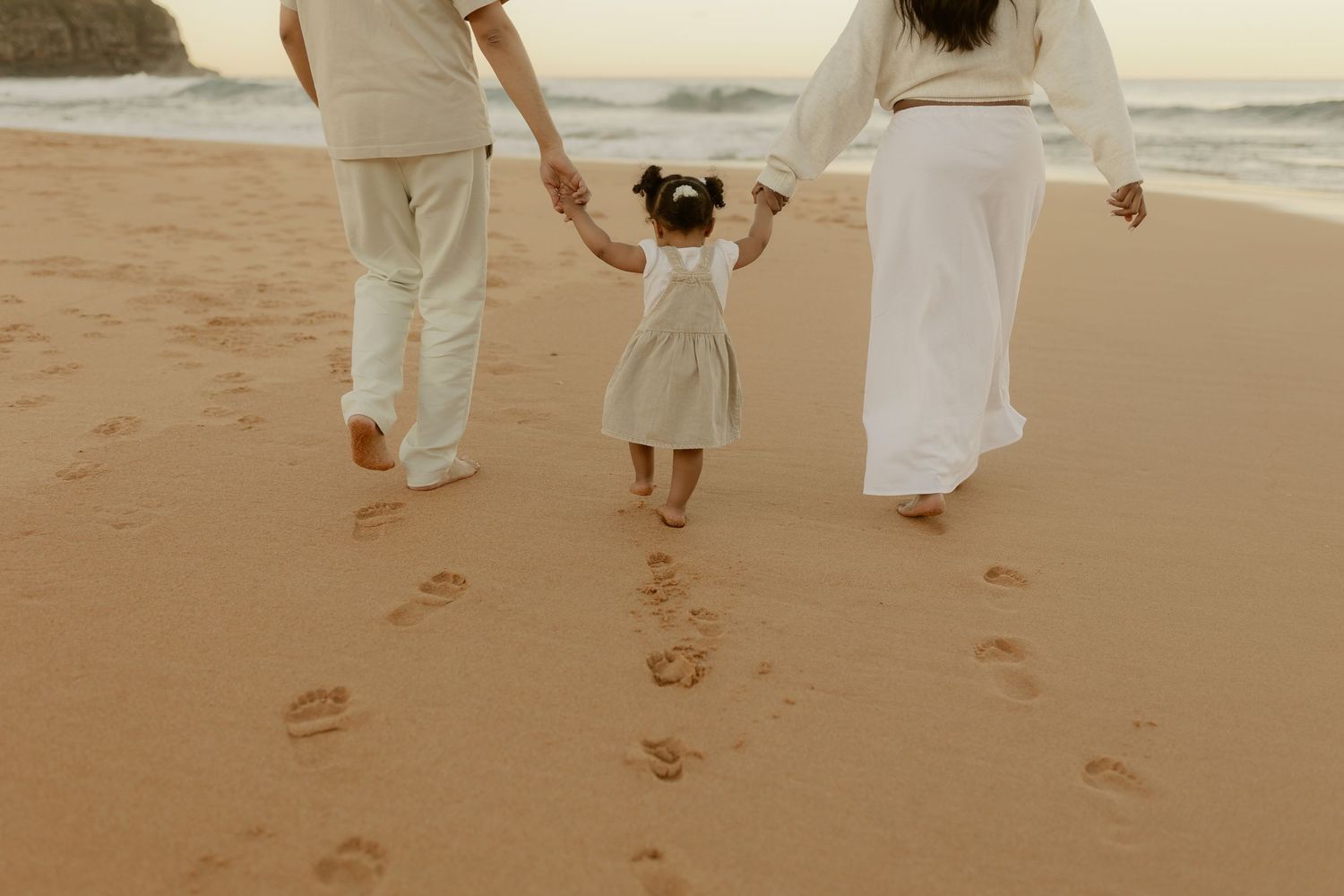 Family of three walking hand-in-hand on sandy beach at sunset, leaving footprints in the sand.