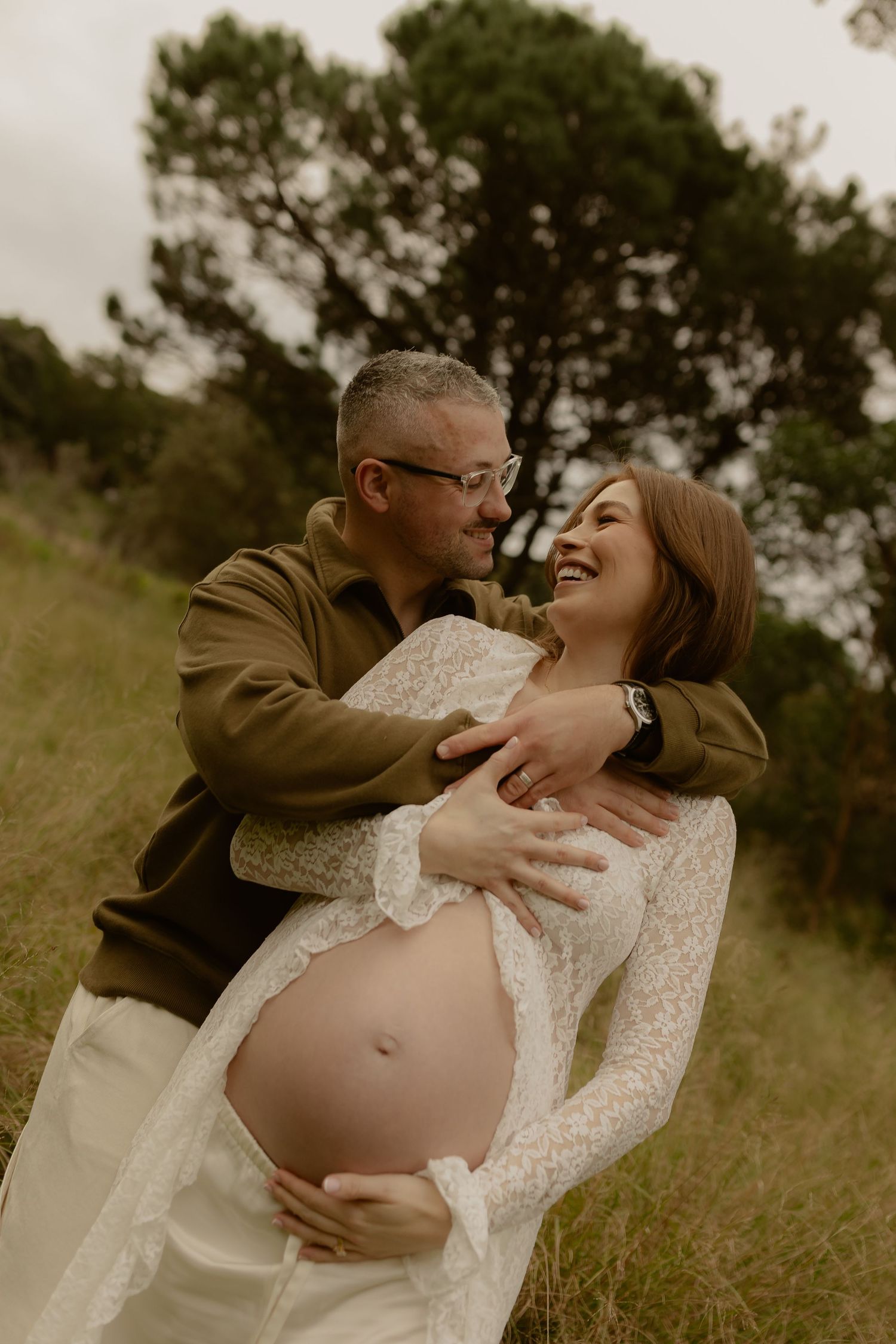 Couple sharing a joyful maternity moment outdoors, man embracing pregnant woman in white lace dress in a green field.