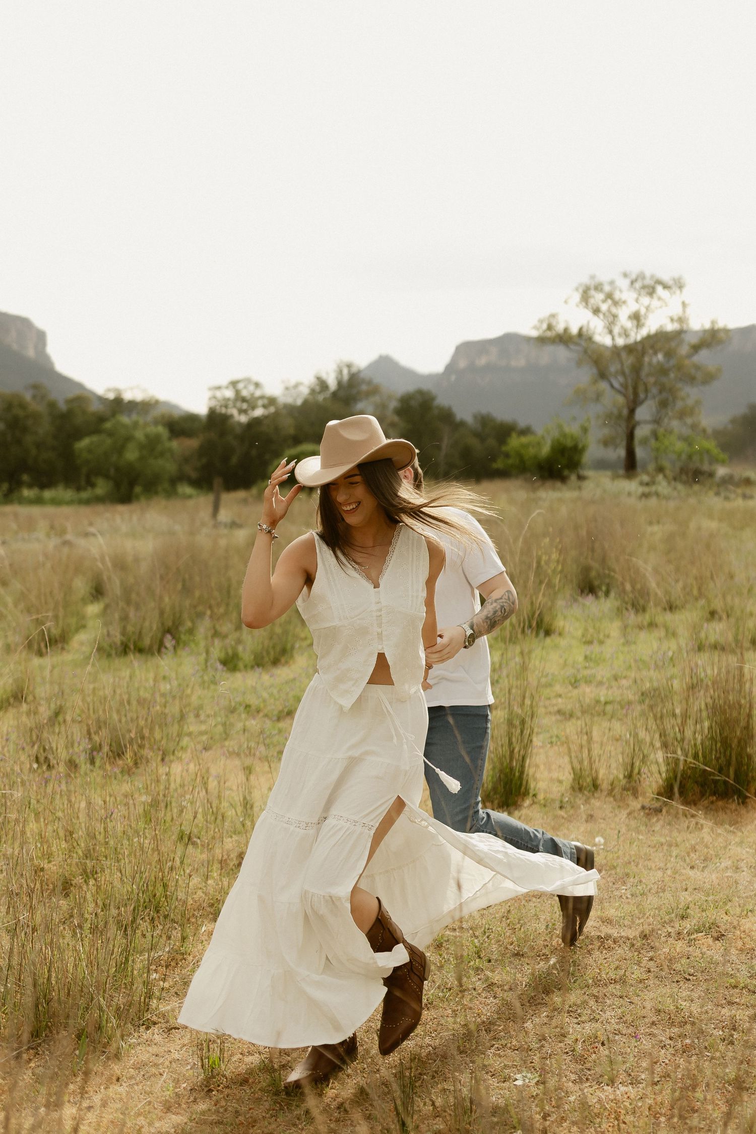 A person in a flowing white dress and cowboy hat dances joyfully in a grassy field.