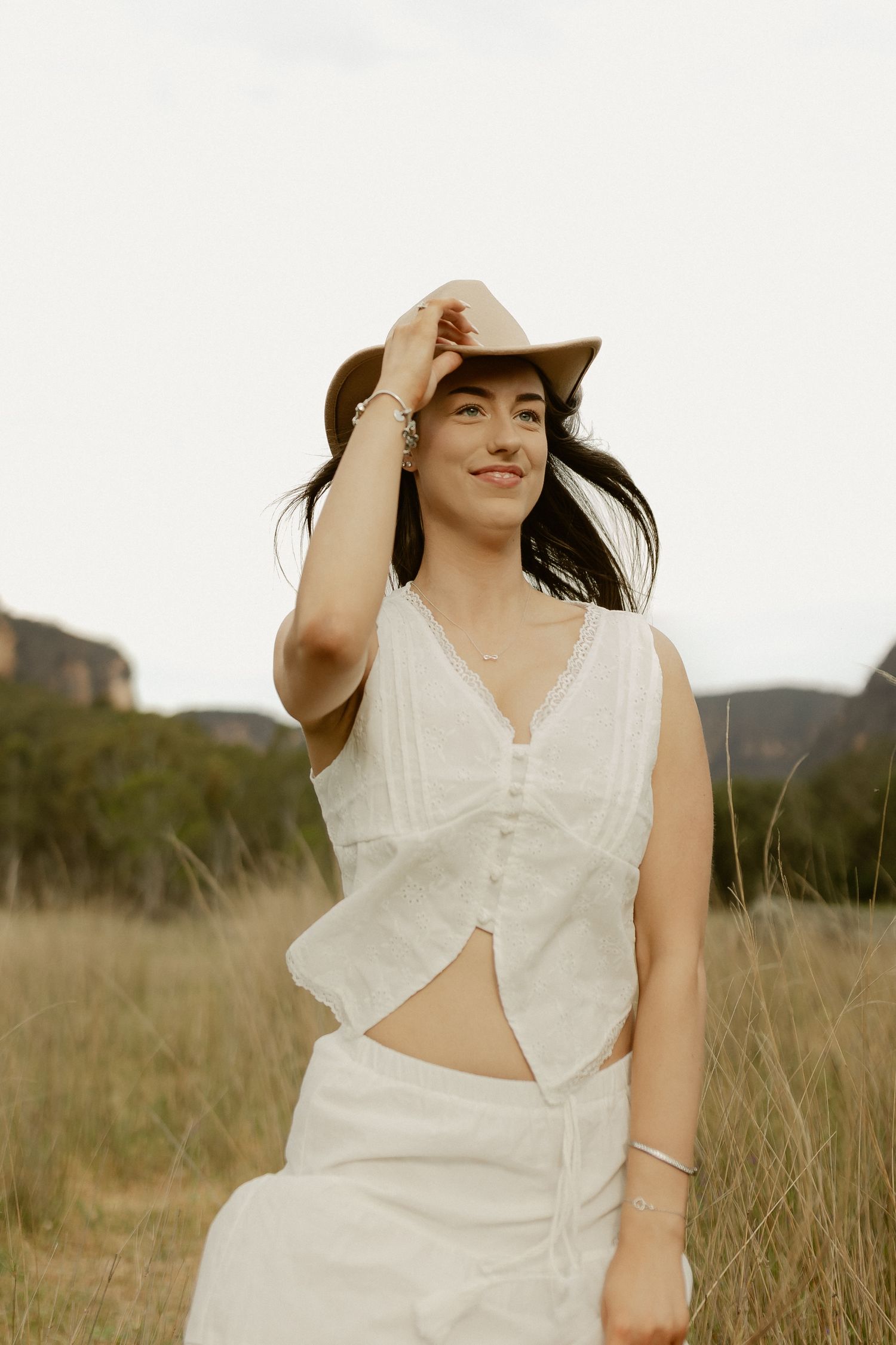 A person wearing a white sleeveless dress and cowboy hat poses in a sunlit field.