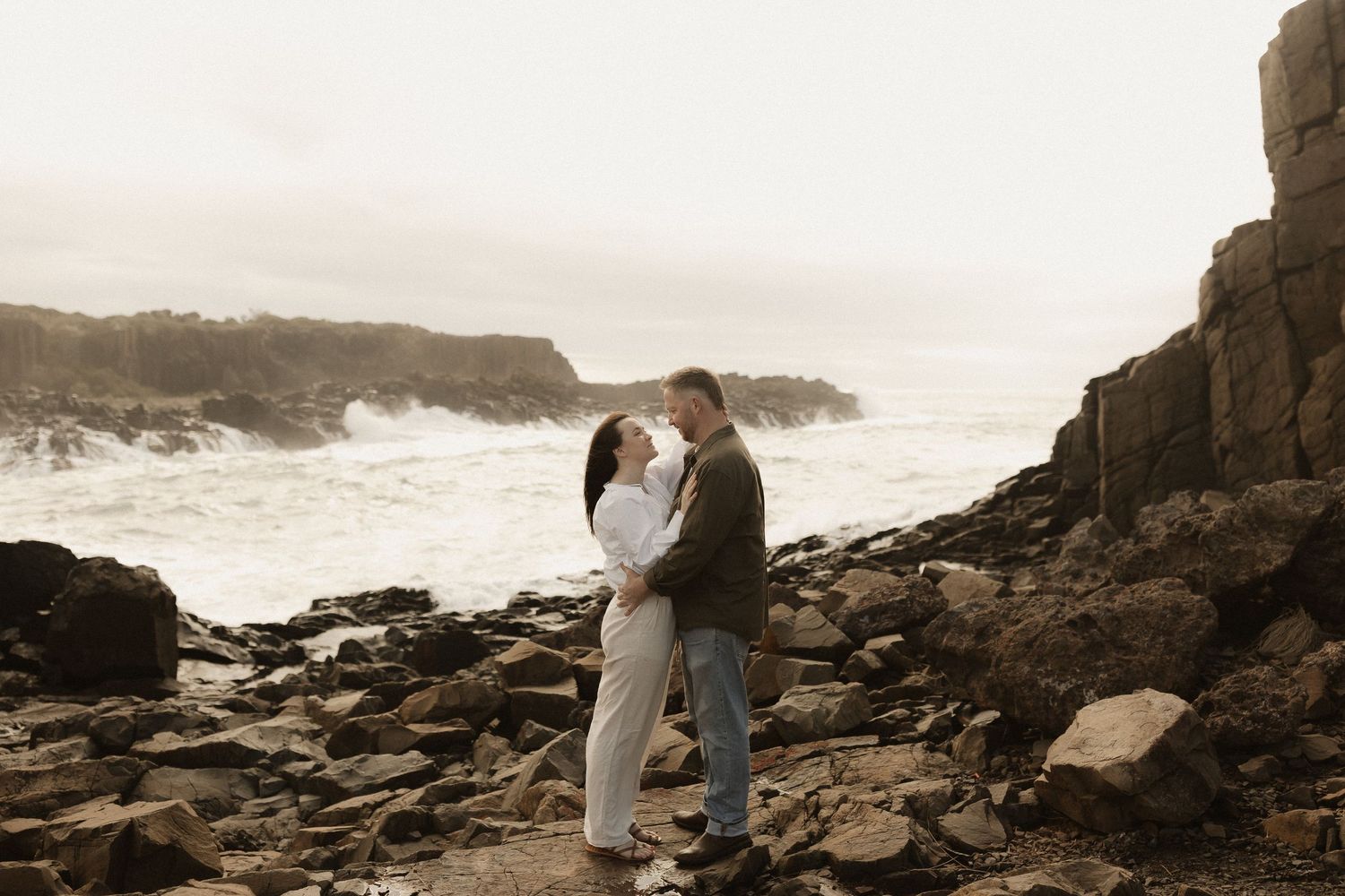 Romantic couple embraces on dramatic rocky shore with crashing waves and cliffs in background.