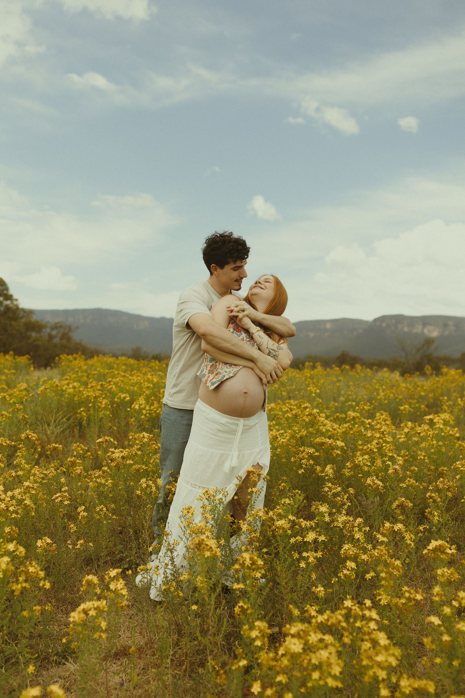 A tender embrace photographed in a field of bright yellow wildflowers with mountains on the horizon.