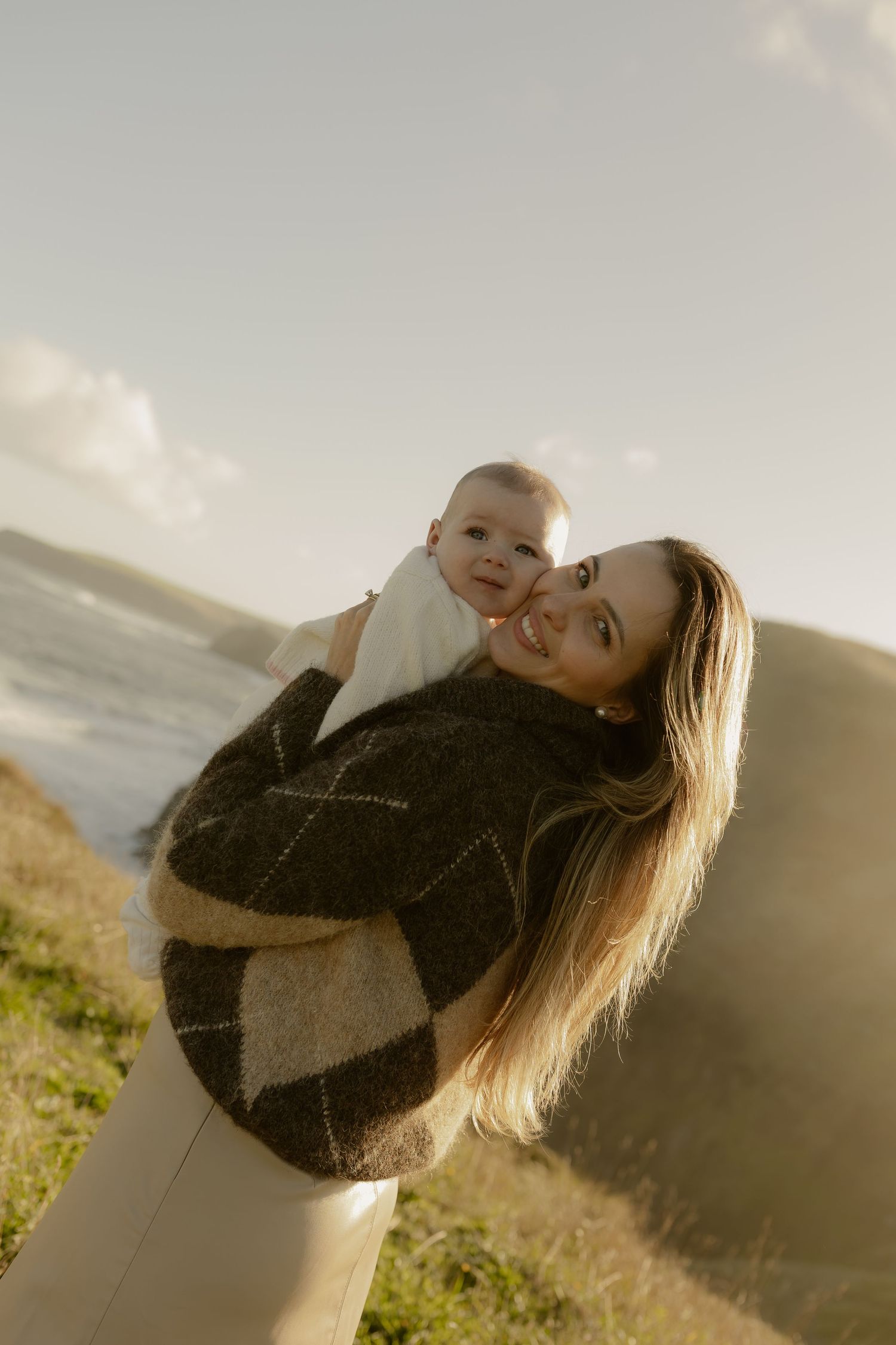 A smiling mother holds her baby outdoors near the ocean, wearing a cozy argyle sweater on a sunny day.