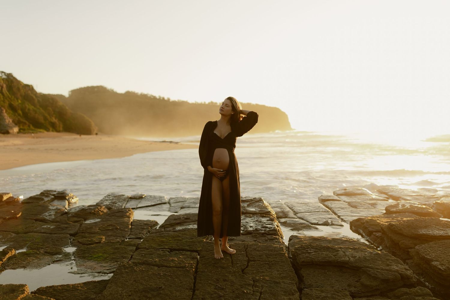 Pregnant woman in black dress standing on rocky beach at golden sunset with ocean waves and cliffs.