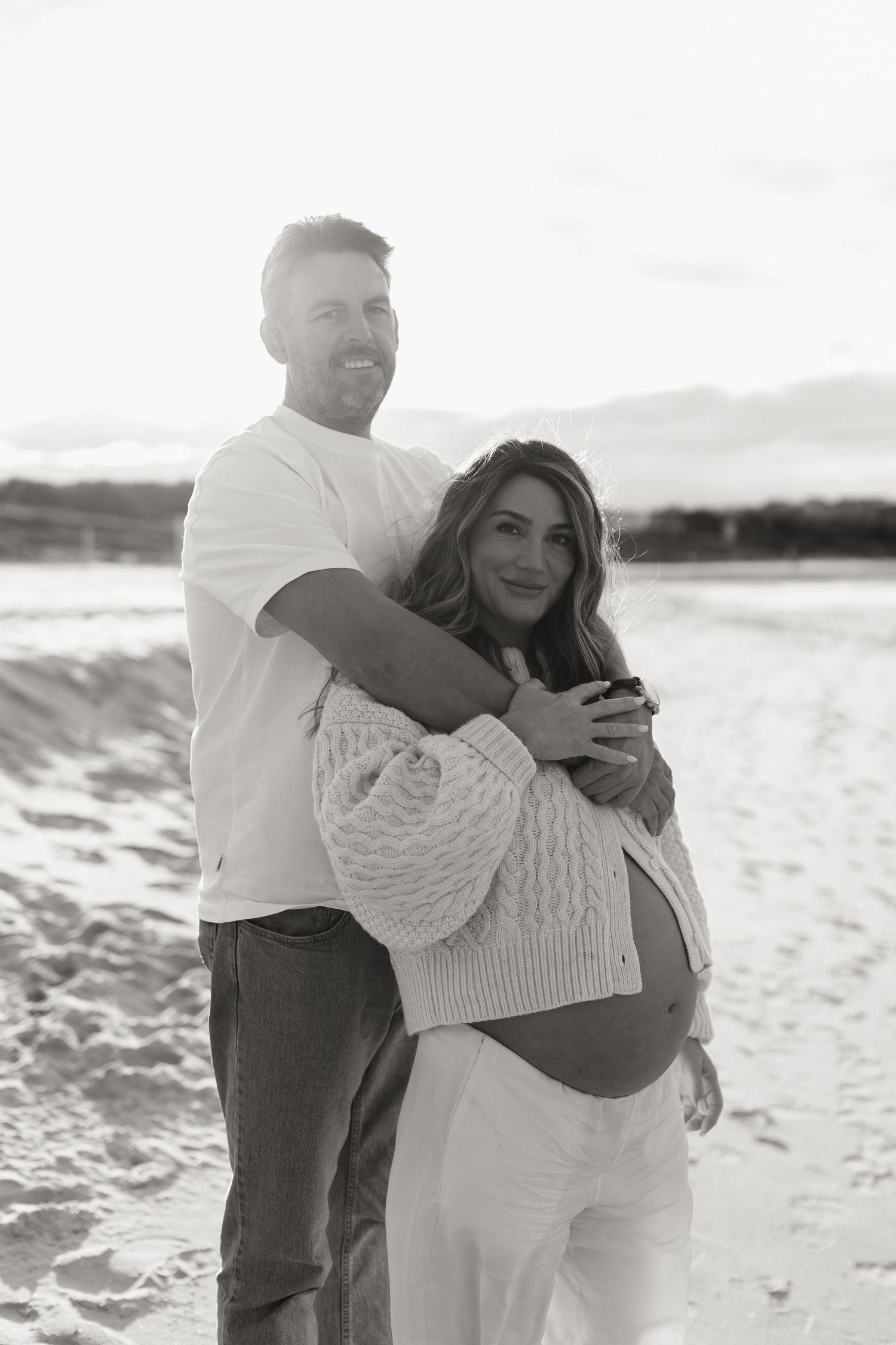 Smiling couple posing on sandy beach, man embracing pregnant woman from behind in black and white maternity photo.