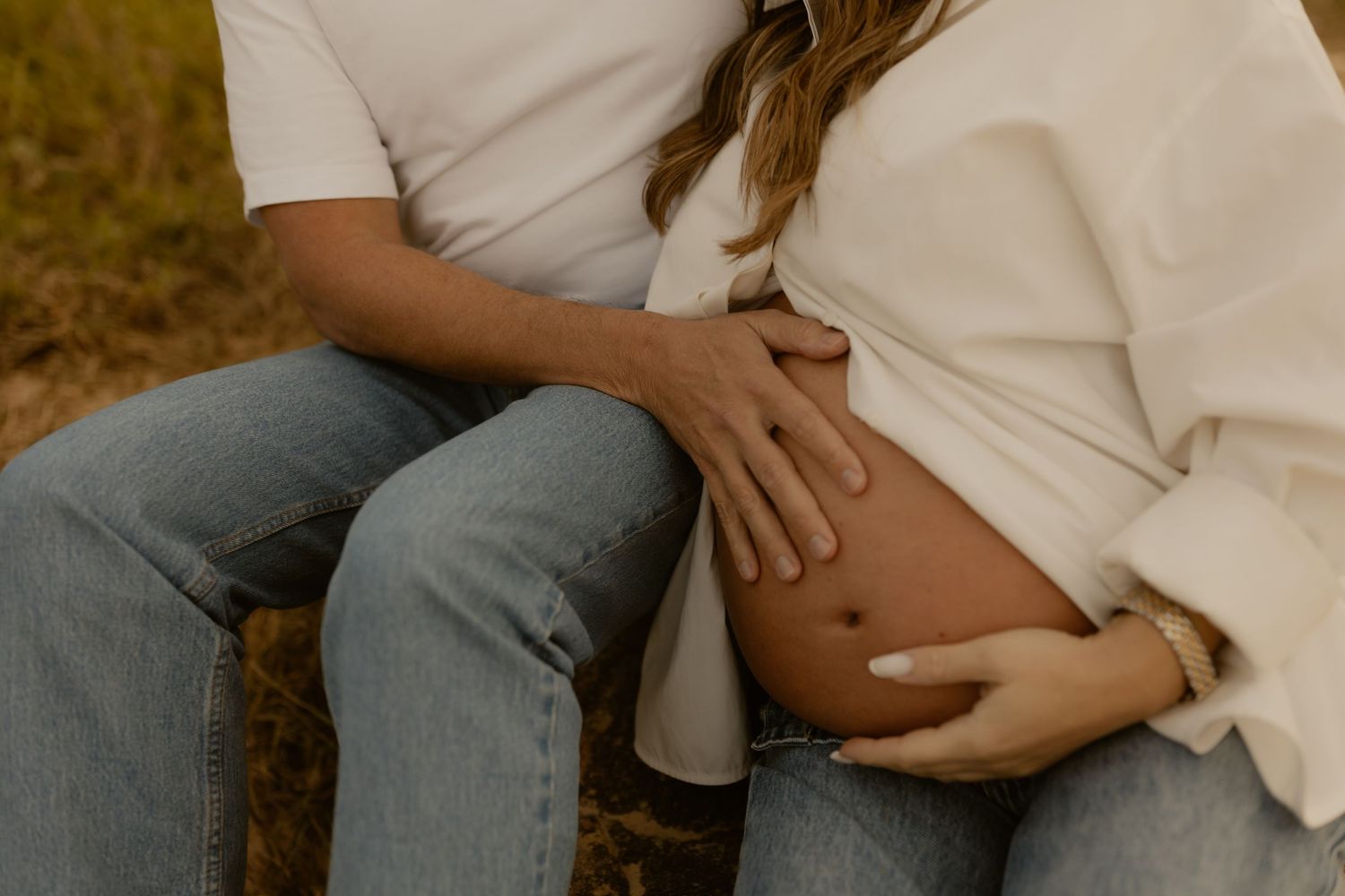 A couple sits together outdoors, both gently cradling a pregnant belly with their hands.