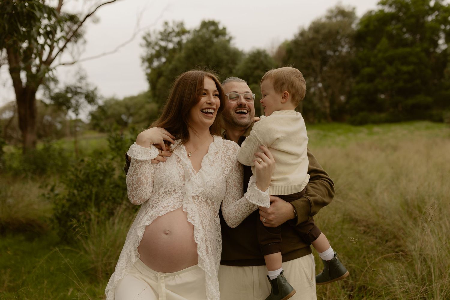 Pregnant woman in lace dress laughing with partner holding toddler outdoors in green field.