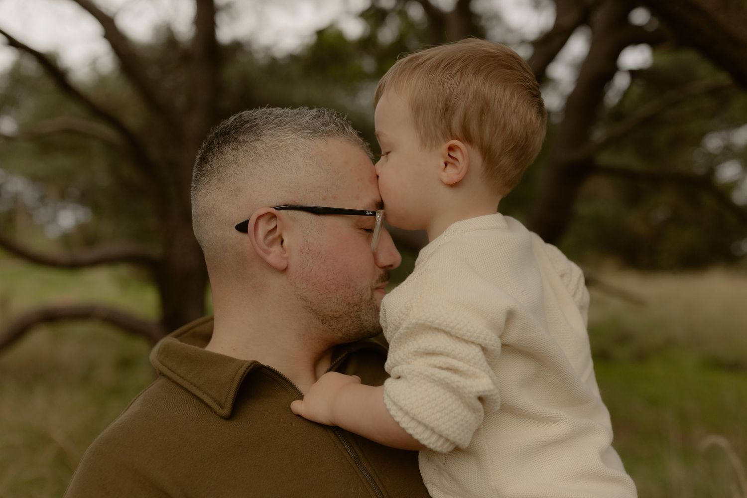 A father and toddler share a sweet nose-to-nose moment outdoors surrounded by trees.