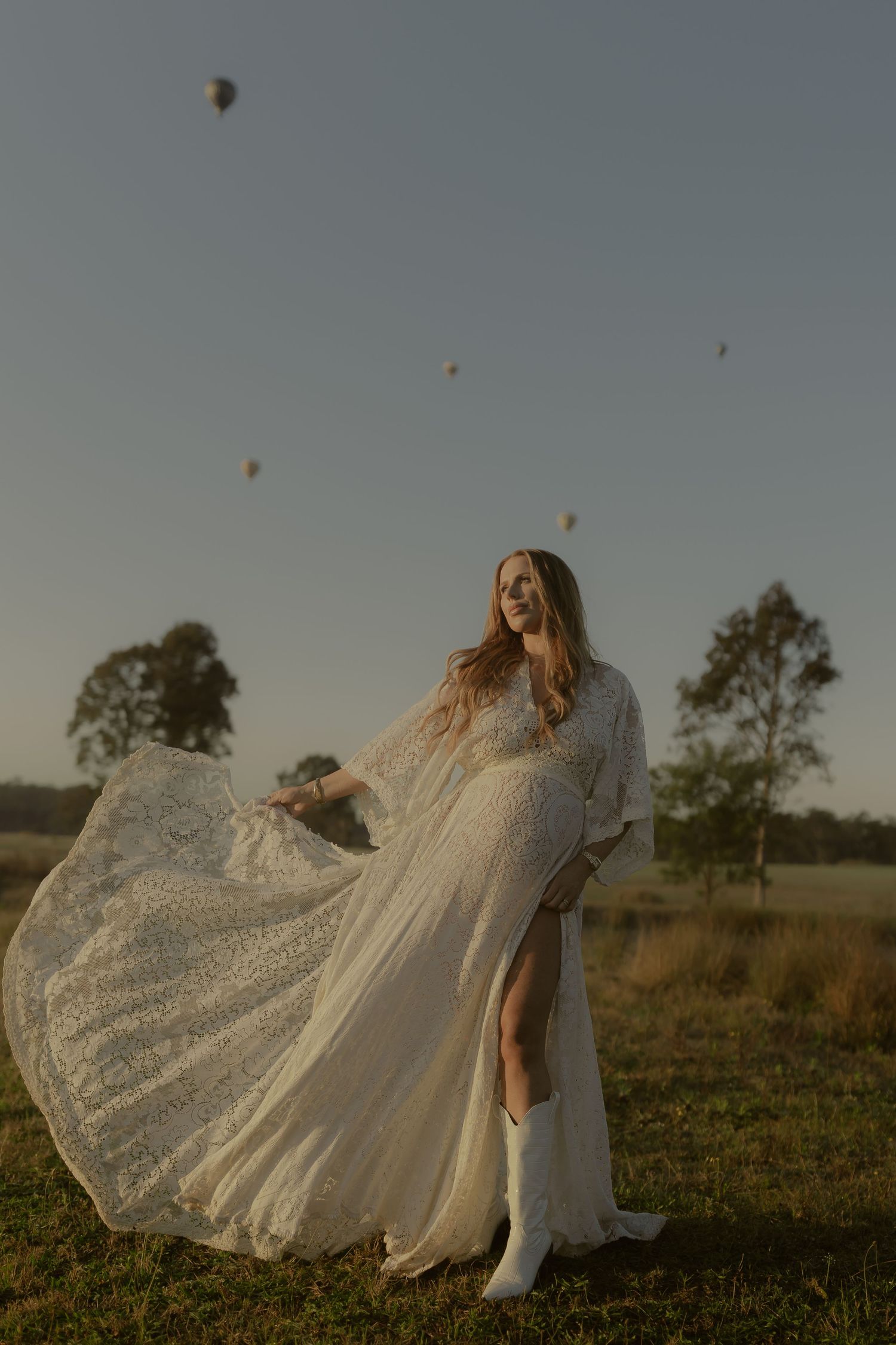 A bride in a flowing white lace gown and boots stands in a field at sunset with hot air balloons in the sky.