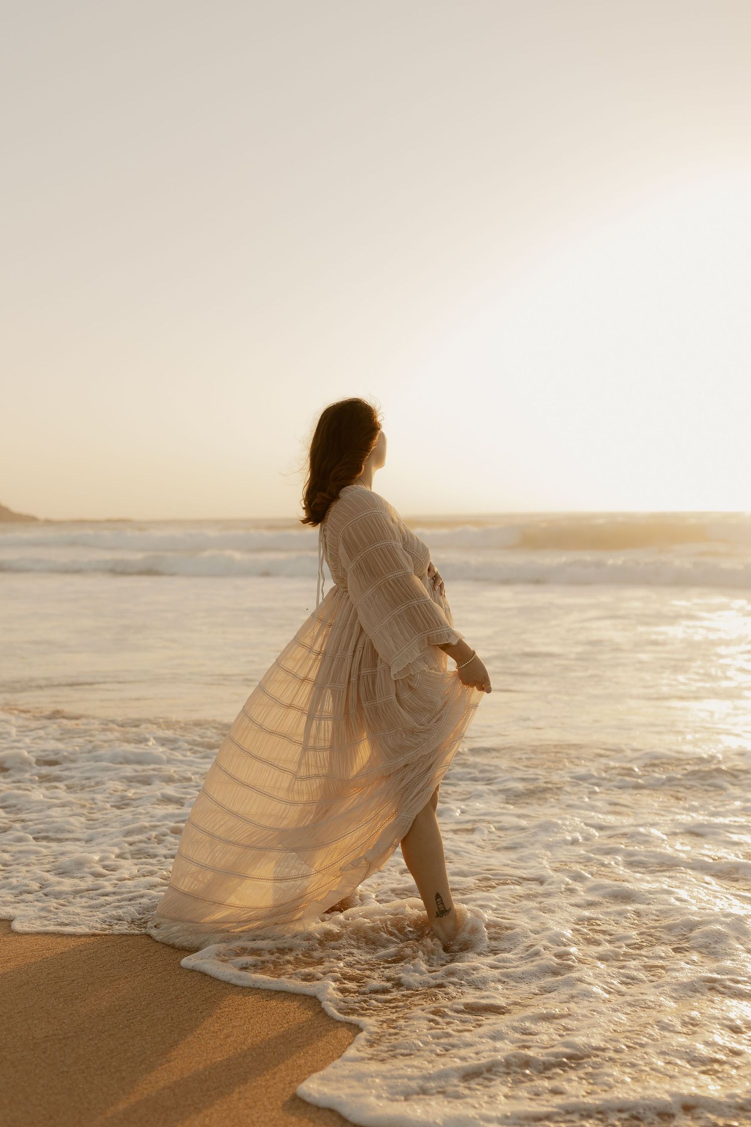 Person in flowing white dress kneels at the ocean's edge during golden hour.
