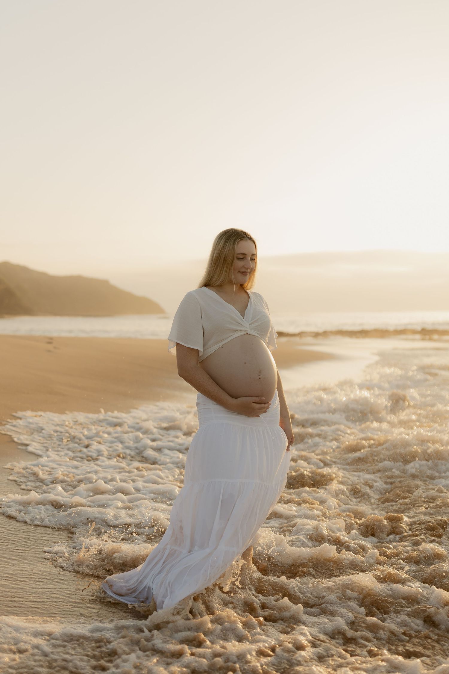 A pregnant woman in a flowing white dress sits in the ocean waves during golden hour.