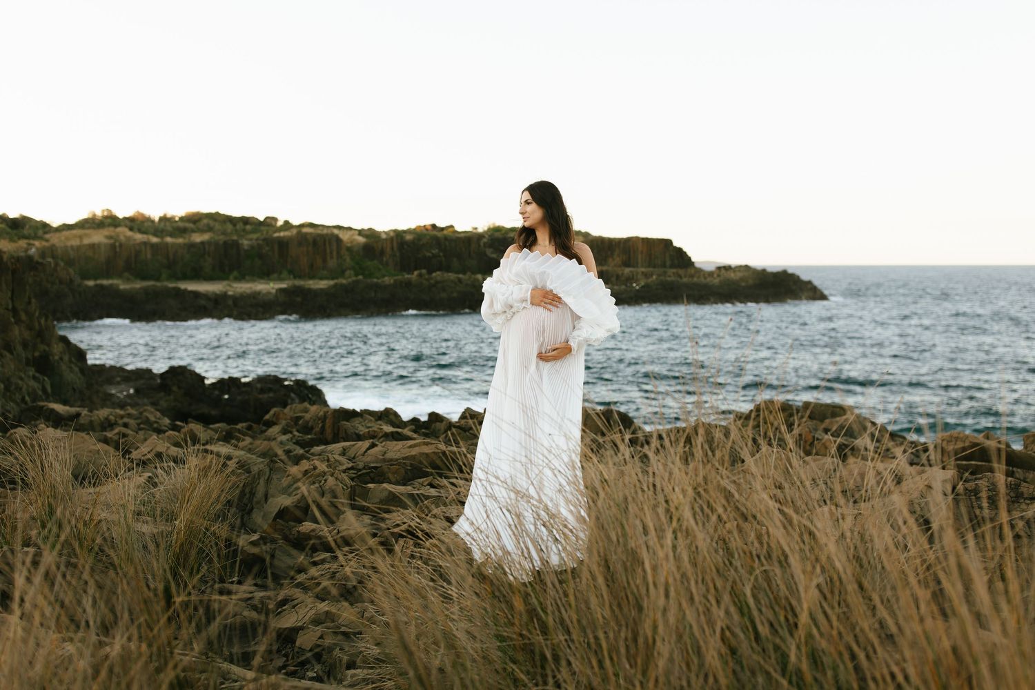 Woman in flowing white dress stands on coastal cliffs overlooking ocean at sunset.