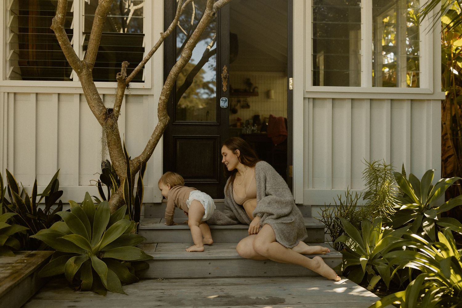 People relax on the front steps of a white porch surrounded by lush tropical plants on a sunny day.