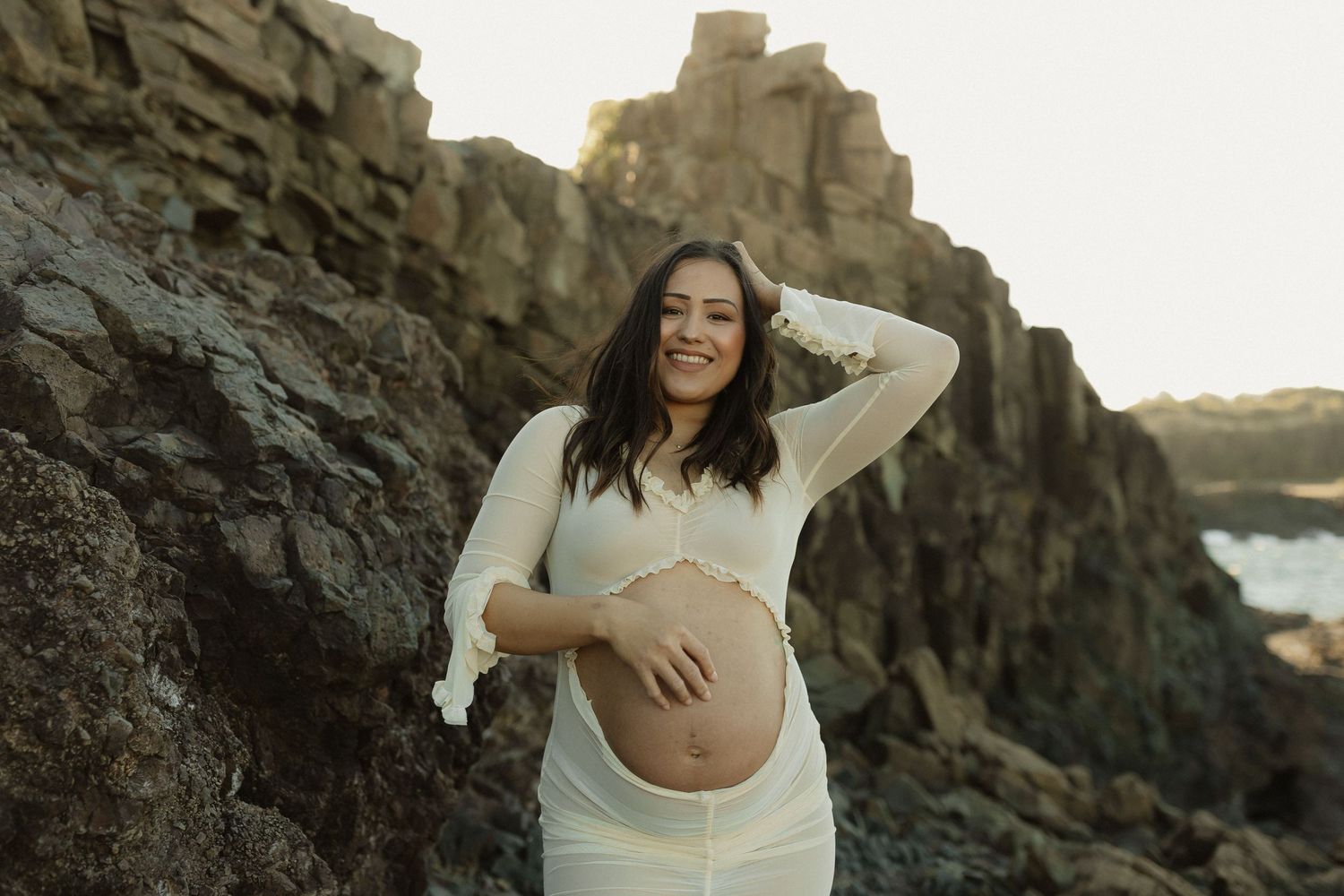 Expectant mother in white attire poses near rocky coastal cliffs during a sunset photoshoot.
