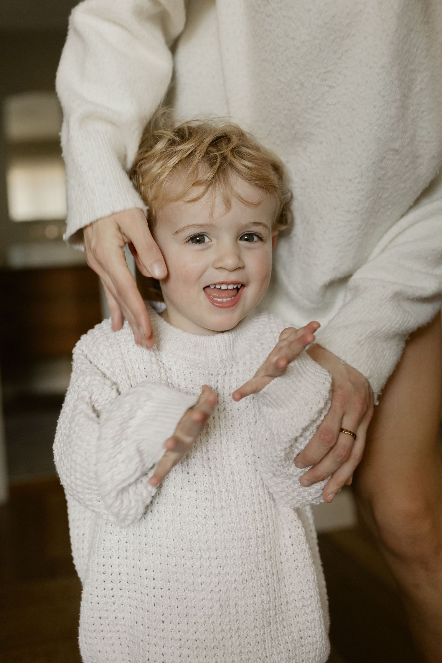 Happy toddler in white knit sweater laughs while being embraced.