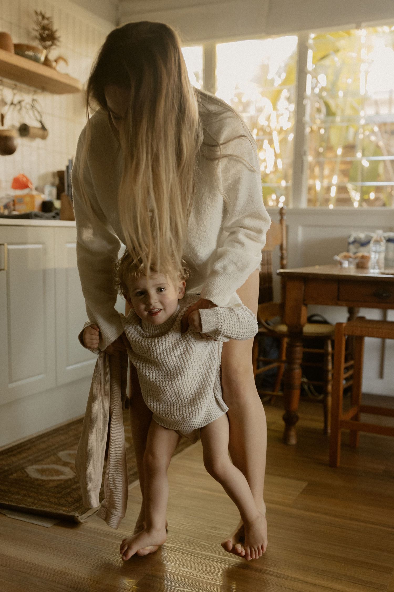 A figure in a white knit sweater leans down while playing on hardwood floors with warm sunlight.