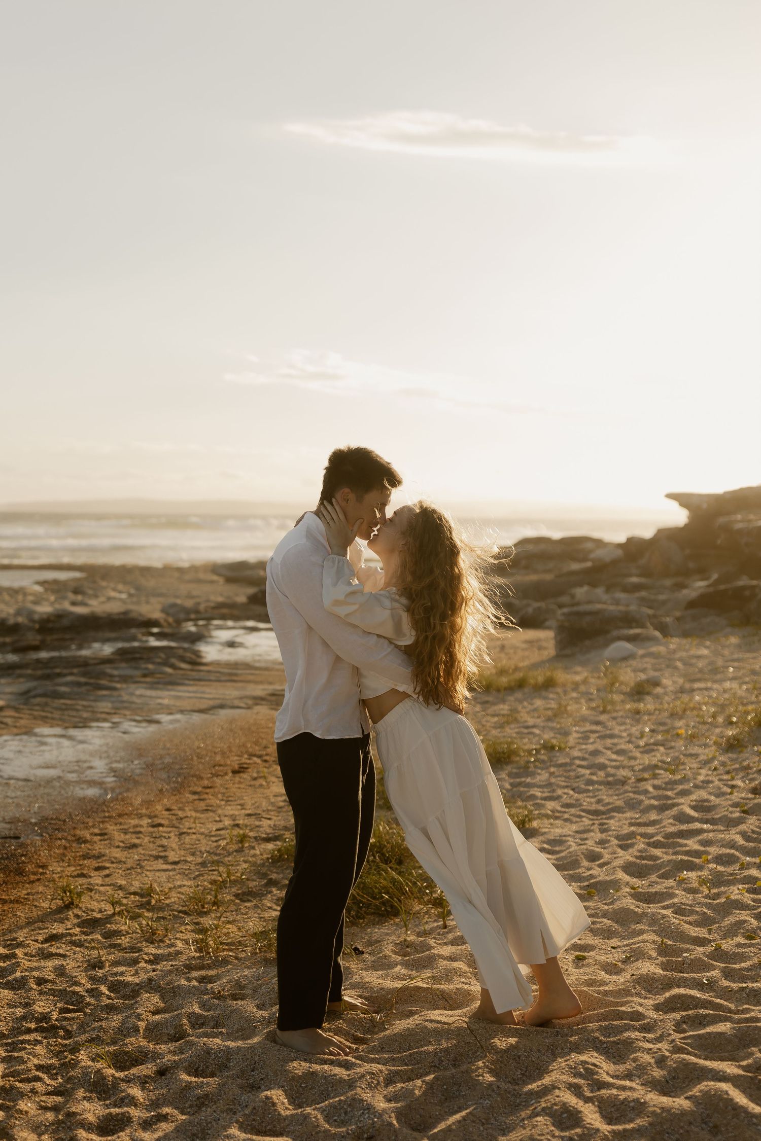 Two people share romantic moment on sandy beach at sunset with ocean in background.