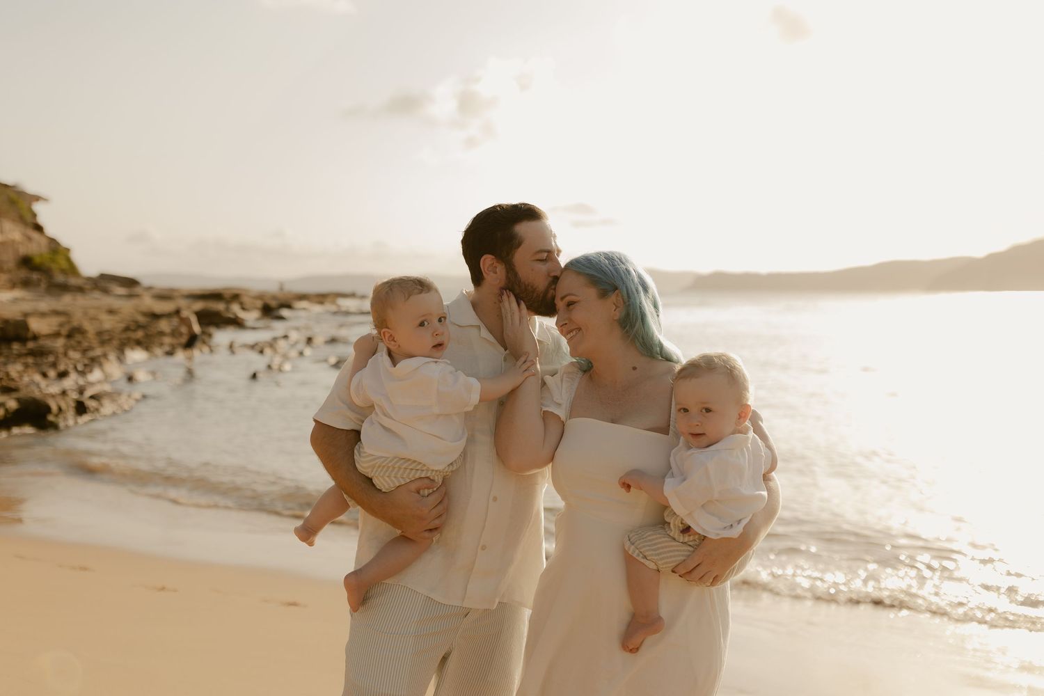 A family wearing white outfits poses together on a sunny beach during golden hour.
