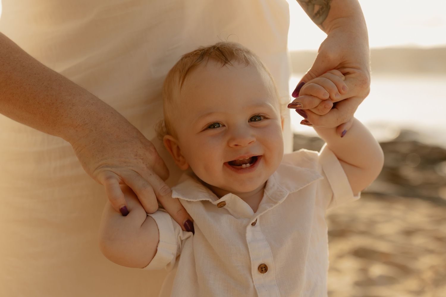 A baby smiles and laughs while being held up in a warm, sunlit setting with soft sepia tones.
