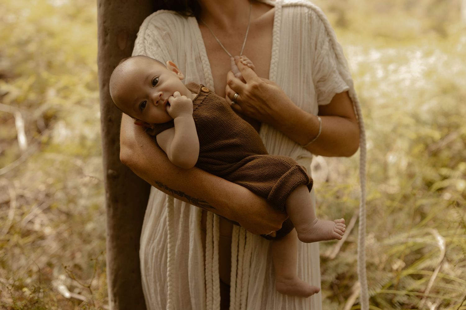 A person in a beige dress holds a small baby while standing in a sunlit field with tall grass.