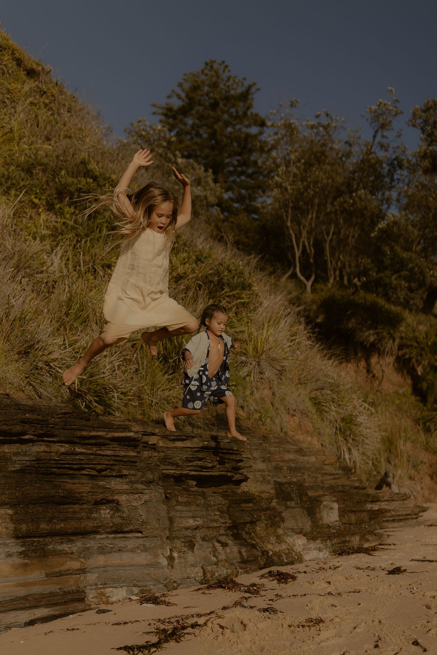 Two people jumping playfully off rocky cliffs at a beach during golden hour.